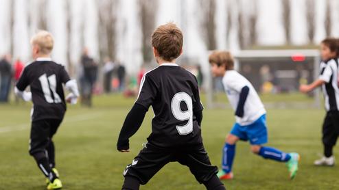 Kinder spielen Fußball auf einem Sportplatz; ein Spieler trägt ein schwarzes Trikot mit der Nummer 9.