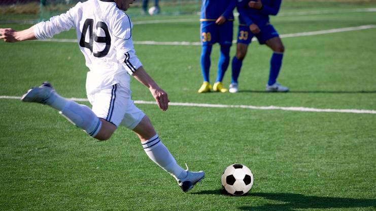 Spieler in weißer Ausrüstung kickt einen Fußball auf einem grünen Kunstrasenplatz, andere Spieler im Hintergrund.
