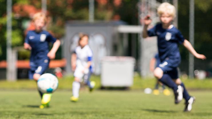 Zwei Kinder in Fußballtrikots rennen auf einem Kunstrasenplatz, ein Ball rollt in der Nähe.