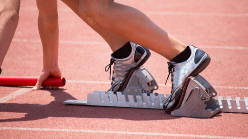 Startposition eines Sprintläufers mit Fuß auf einem Startblock auf einer roten Laufbahn, Hand hält einen Staffelstab.