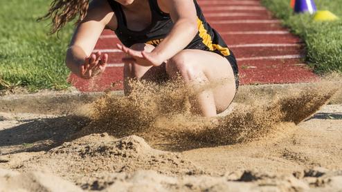 Athletin springt in einen Sandgruben, während Sand aufwirbelt. Grüße des Sprungbretts sind im Hintergrund sichtbar.