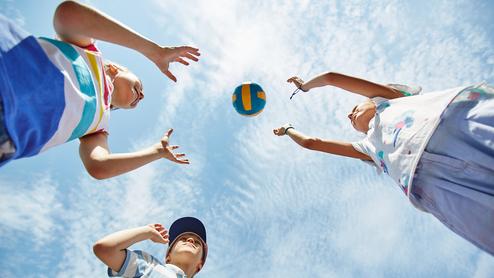 Drei Kinder werfen einen bunten Volleyball in den Himmel vor einem blauen Himmel mit einigen Wolken.